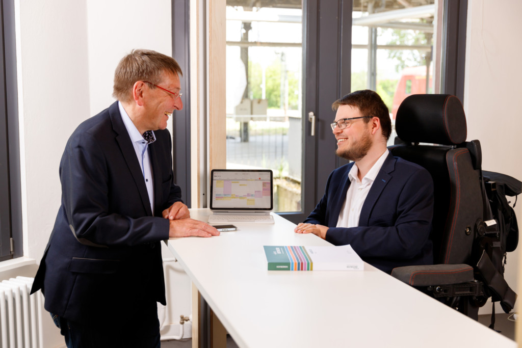 Ulrich Watermann steht gemeinsam mit Constantin Grosch an einem Bürotisch. Vor sich ein iPad und Buch.