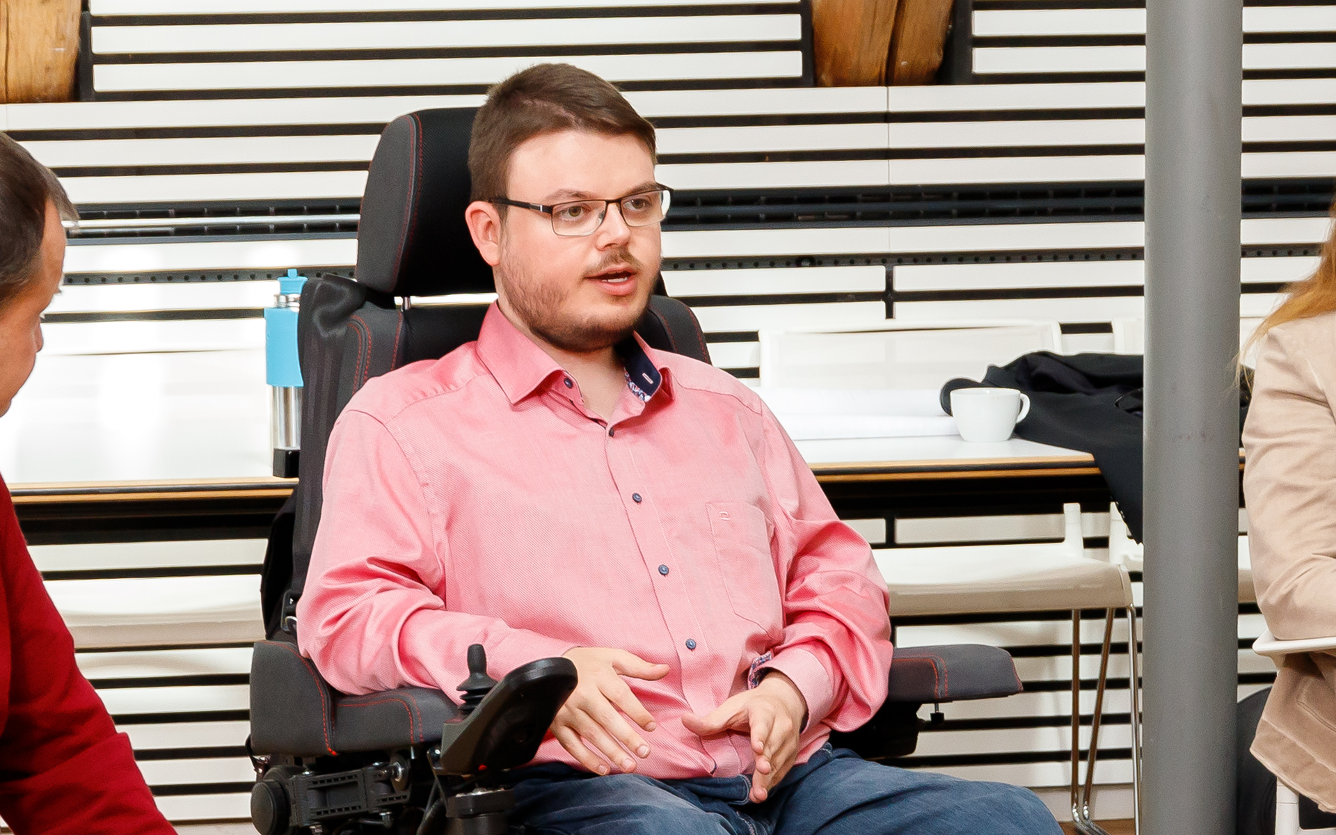 Constantin Grosch sitzt mit rotem Hemd im Rollstuhl und gestikuliert. Links und rechts sieht man Menschen sitzen.