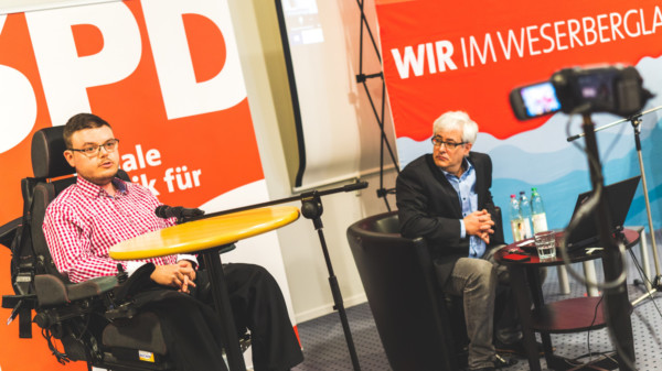 Constantin Grosch steht an einem Stehtisch. Vor ihm eine Kamera und ein Mikrofon. Hinter ihm ein SPD Banner. Rechts neben ihm sitzt Matthias Gombach in einem Sofa und blickt zu ihm.